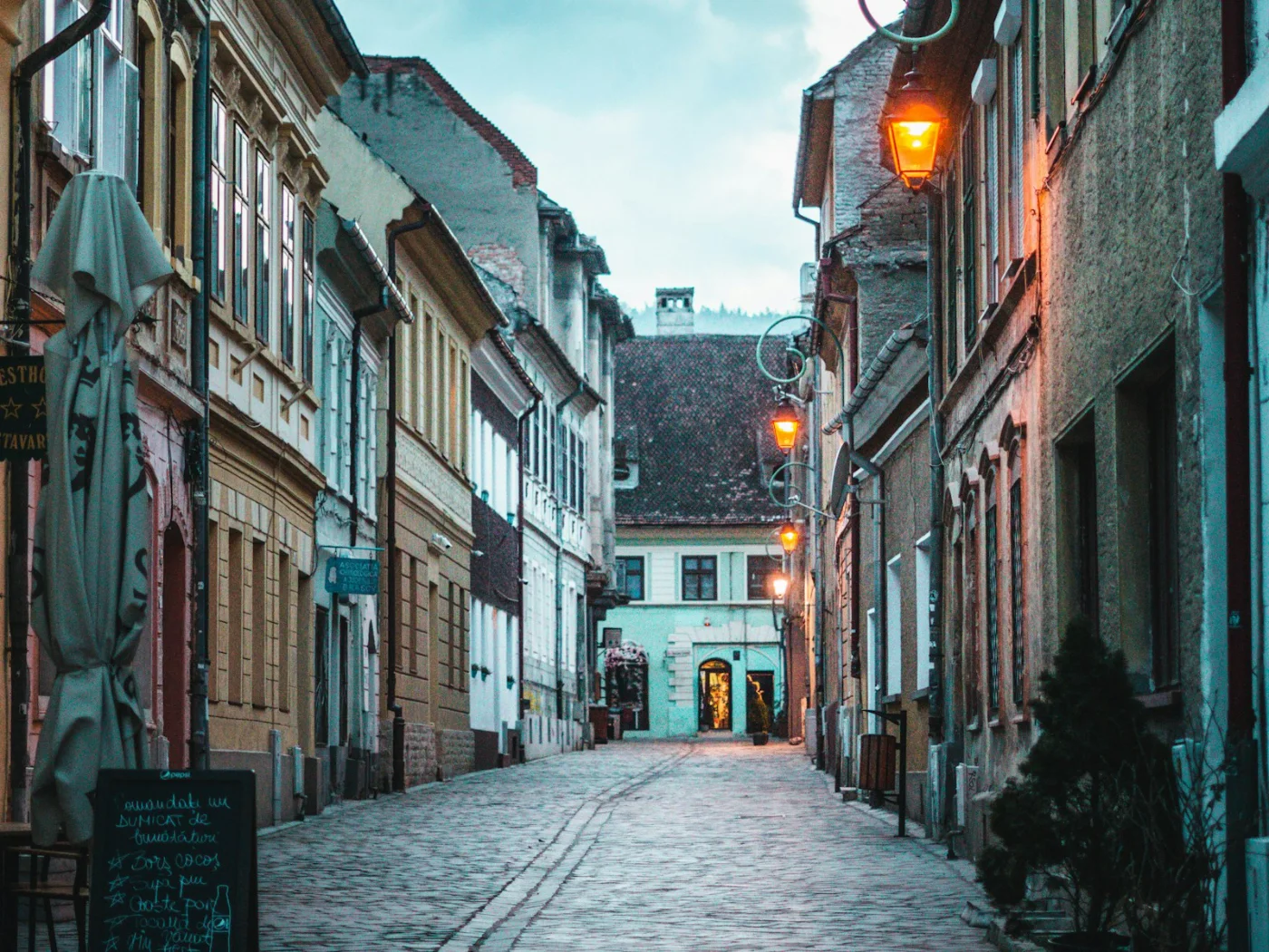 A cobbled street in the old town of Brașov at dusk, lamps lit, with a guesthouse sign and a chalkboard menu outside a doorway